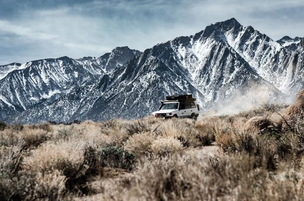 White Misubishi Montero drives through brush with snowy mountains behind.